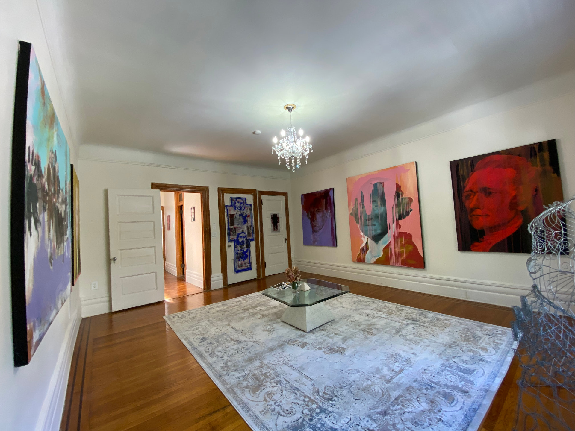 Gallery room with large expressive painted portraits of American founding figures above a glass coffee table, with a wire sculpture in the foreground