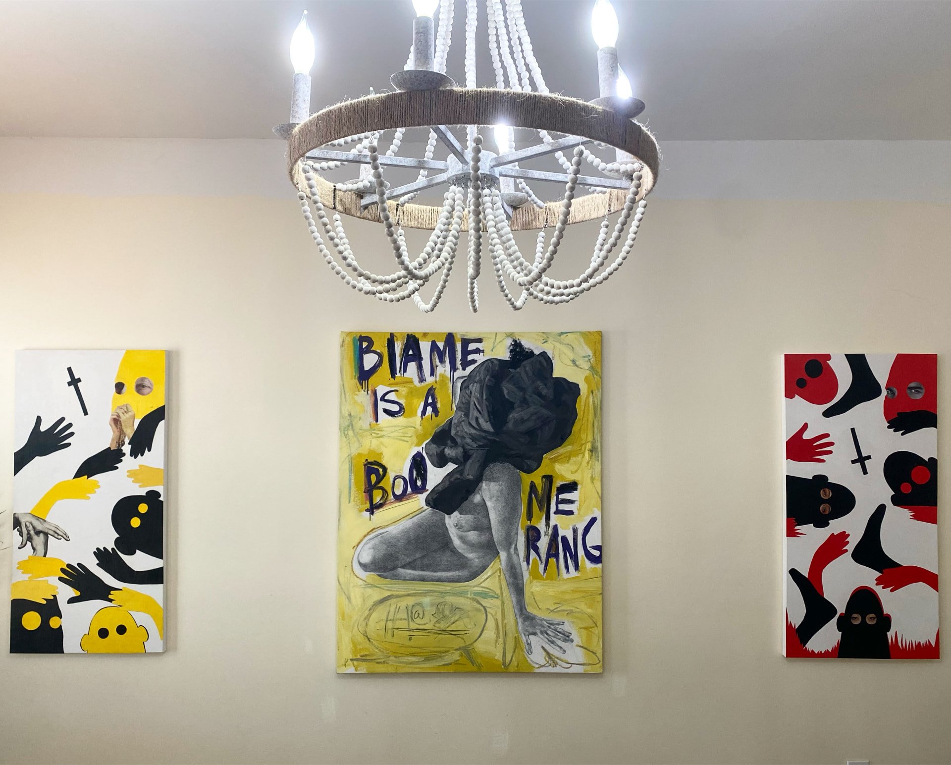 Yellow painting reading Biame is a Boomerang with a kneeling graphite figure, flanked by two figurative works with symbolic hands, beneath a beaded chandelier