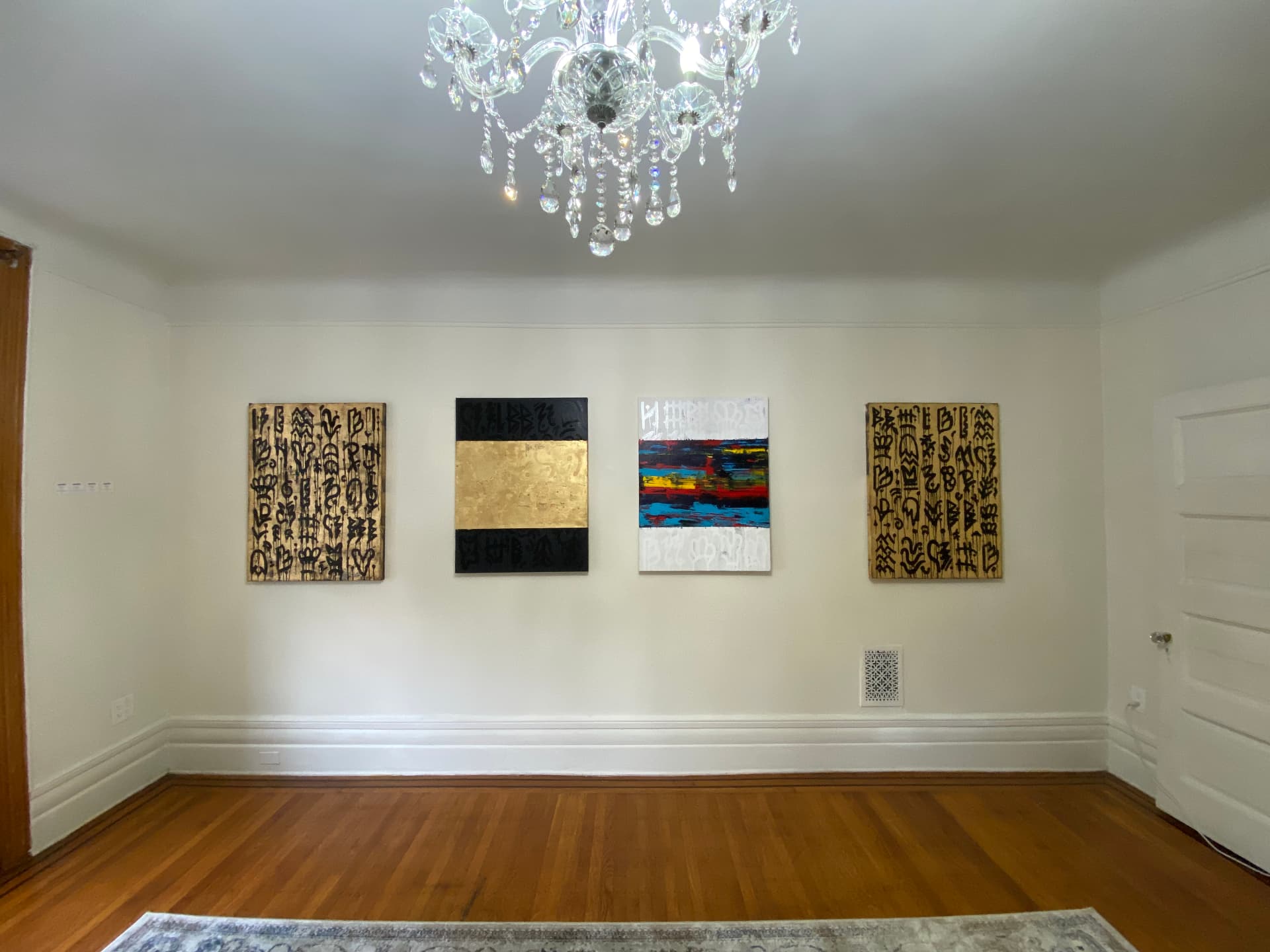 Installation view — four works by Braxton Fuller, including two gold-leaf glyph canvases, a gold-and-black band painting, and a multicolored abstract, hung in an elegant dining room beneath a crystal chandelier