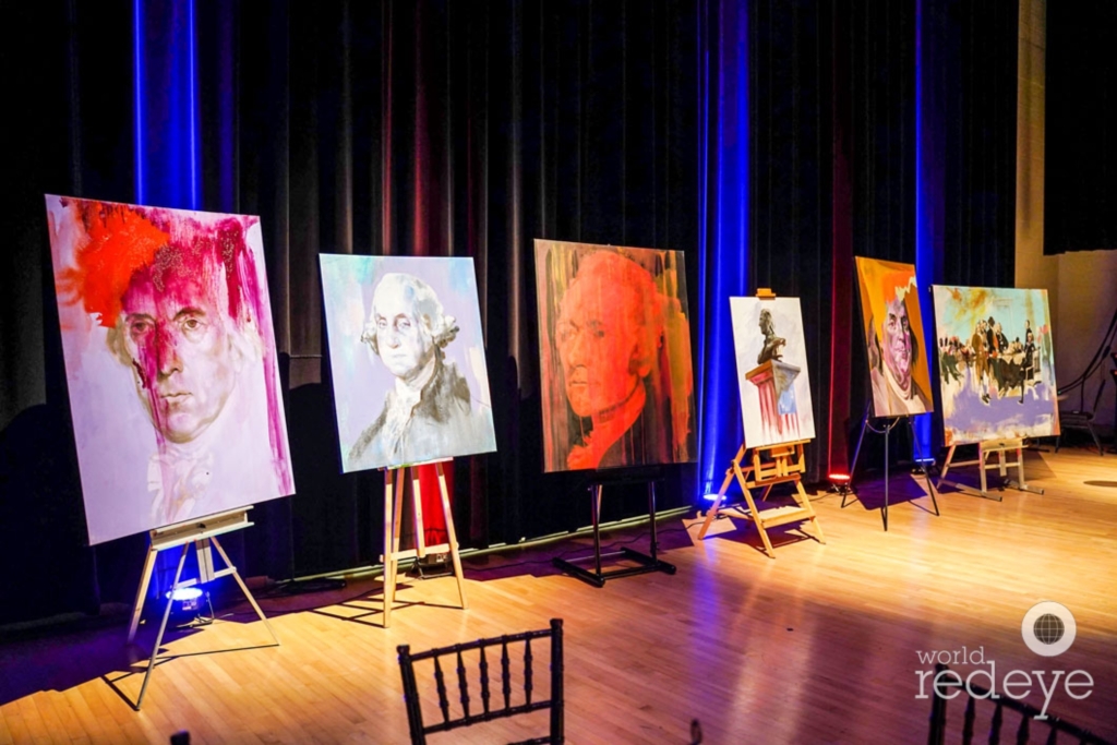 Stage of the Hamilton exhibition at the Adrienne Arsht Center, paintings of American founding figures displayed on easels under theatrical blue lighting